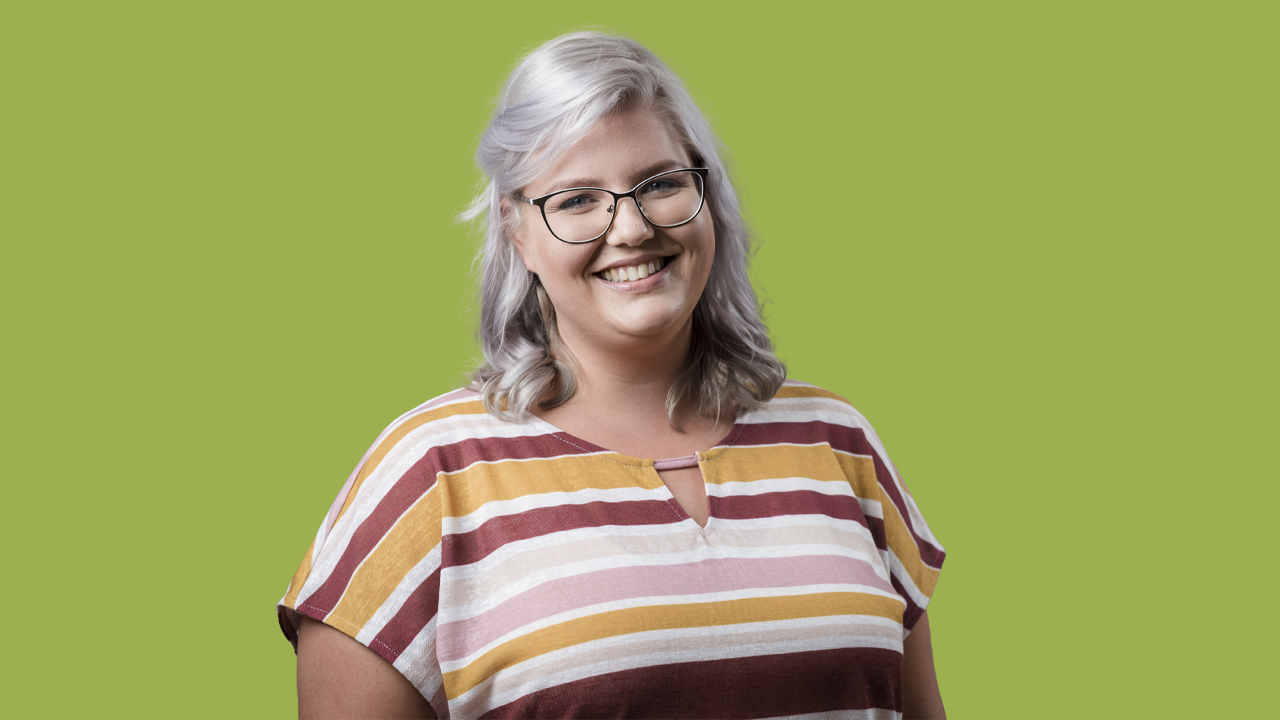 Eine junge Frau mit weißblonden Haaren und Brille steht vor grünem Hintergrund. Ihr Lächeln strahlt Wärme und Offenheit aus.